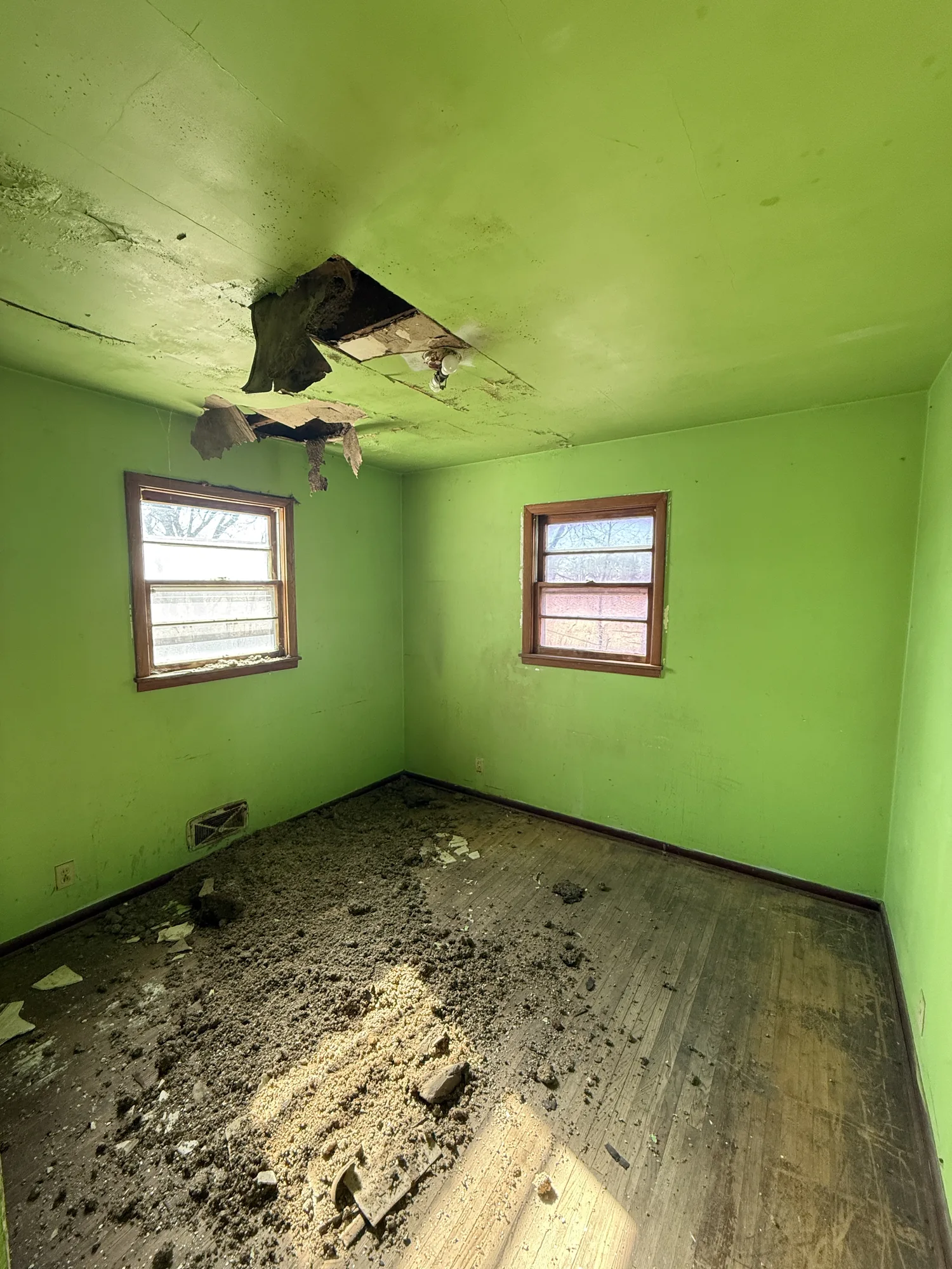Storm-damaged bedroom with a collapsed ceiling section, fallen insulation and debris across the floor, and water-stained green walls