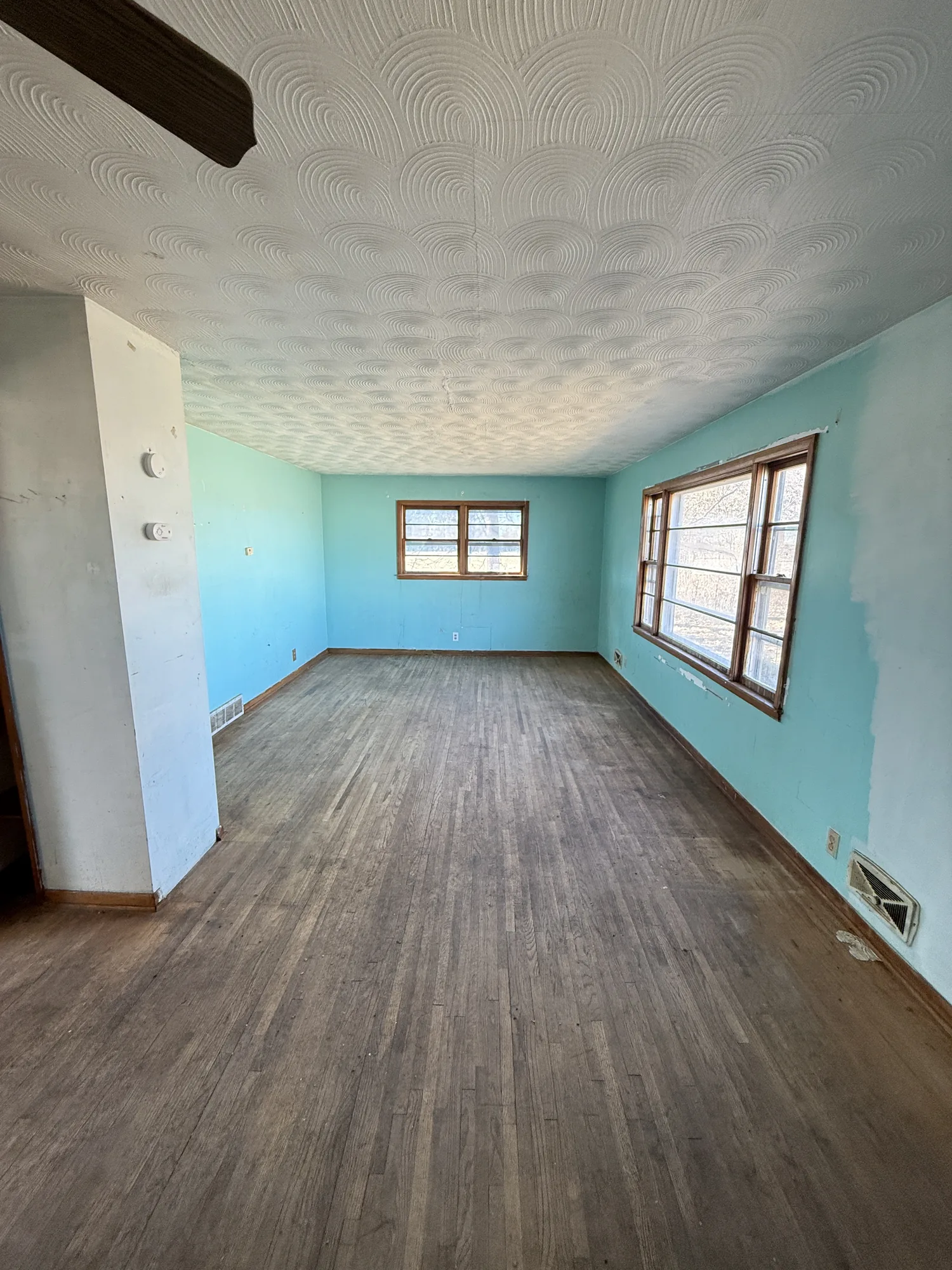 Gutted farmhouse living room with damaged wood flooring, peeling teal paint, and a cracked swirl-textured ceiling