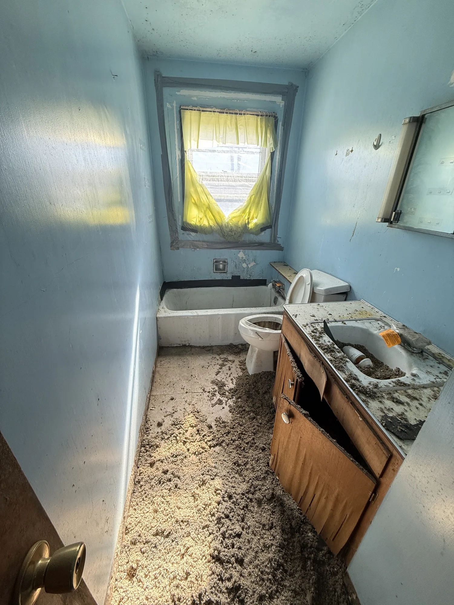 Water-damaged bathroom in the Austin farmhouse with a debris-covered floor, peeling blue paint, and a worn tub and surround