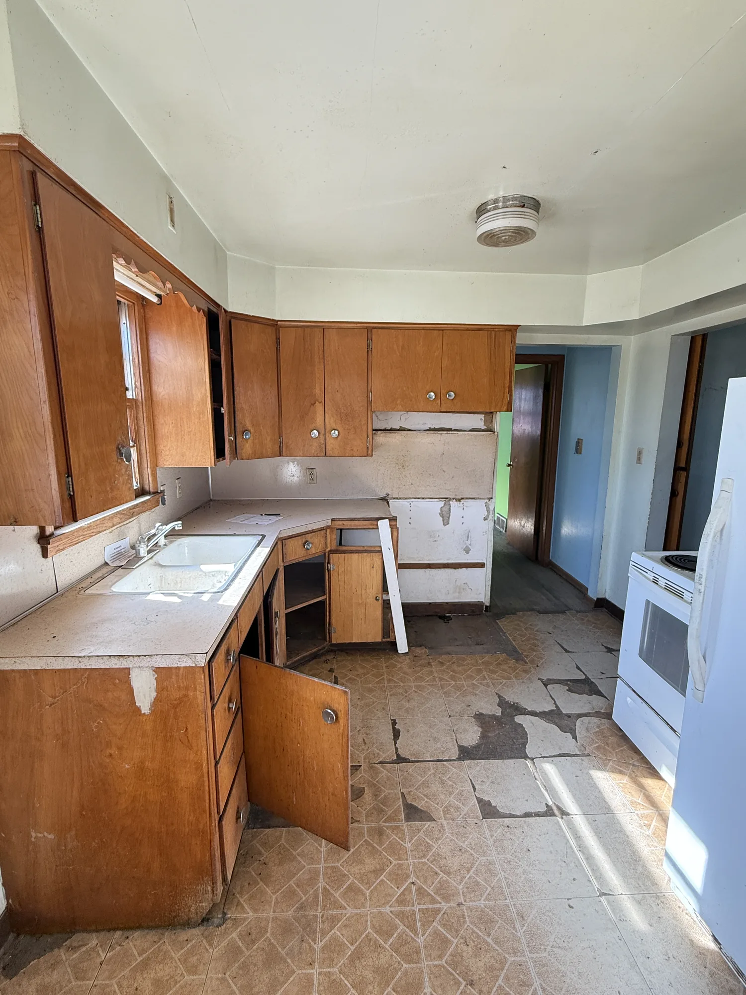 Damaged farmhouse kitchen in Austin, MN with worn wood cabinets, missing doors, peeling flooring, and water-stained walls