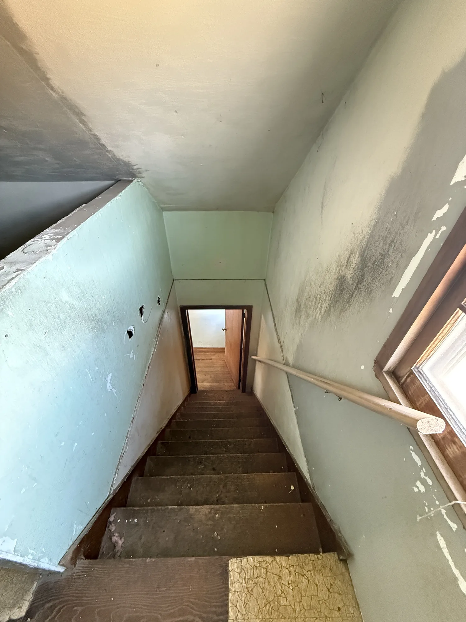 Damaged farmhouse stairwell with peeling paint, worn wooden steps, and debris on the treads